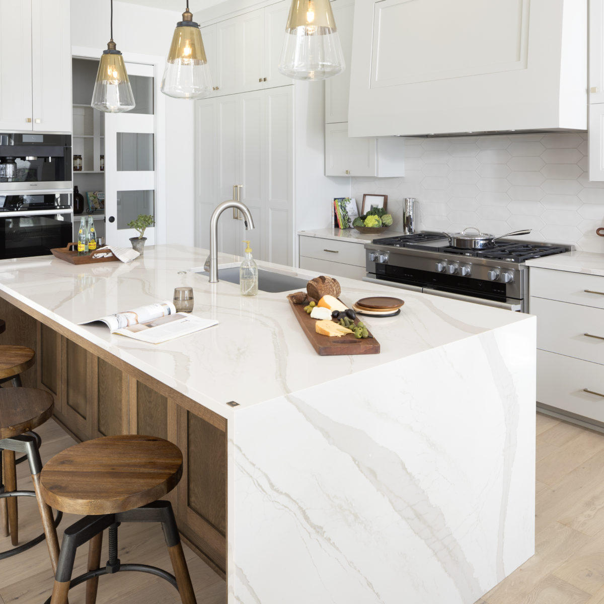 Brittanicca Warm Cambria Quartz Kitchen Countertops and Island with Waterfall Panels Brown Cabinets Pendant Lighting